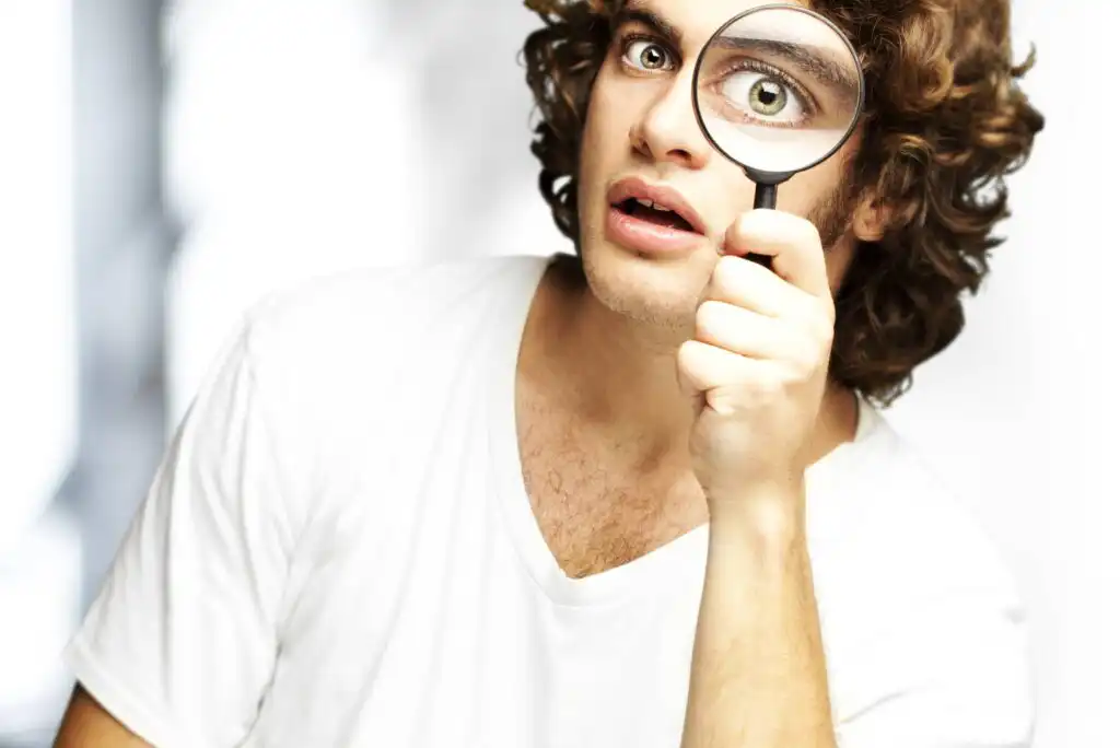 A young man with curly hair wearing a white shirt holds a magnifying glass in front of one eye, making his eye appear much larger. He has a surprised expression. The background is bright and blurred.