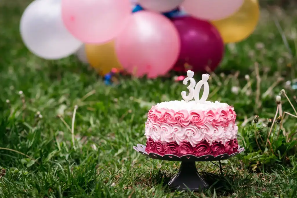 A pink and white frosted cake with a 30 candle sits on a stand outdoors on grass. Blurred colorful balloons are in the background, suggesting a 30th birthday celebration.