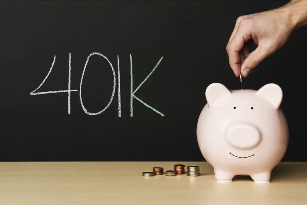 A hand places a coin into a pink piggy bank on a table, with stacks of coins nearby and 401K written in large letters on a blackboard in the background.
