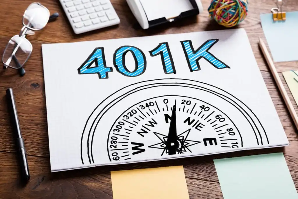 A notebook with 401K written in large blue letters above a compass graphic sits on a wooden desk, surrounded by office supplies like a keyboard, pen, sticky notes, and a rubber band ball.