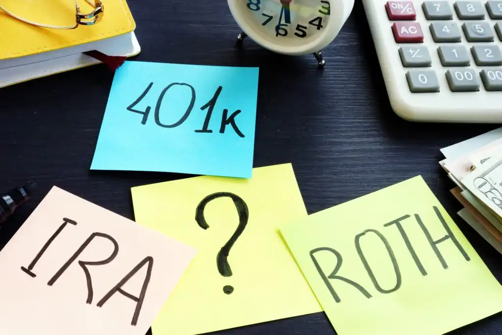 Sticky notes labeled 401k, IRA, ROTH, and one with a question mark are on a desk with a calculator, clock, notebook, cash, and glasses, suggesting decisions about retirement savings options.