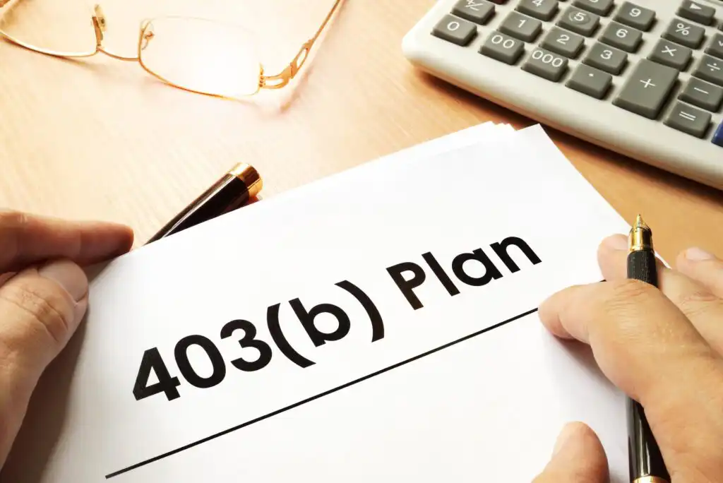 A person holding a document labeled 403(b) Plan on a desk, with a pen, glasses, and a calculator nearby.