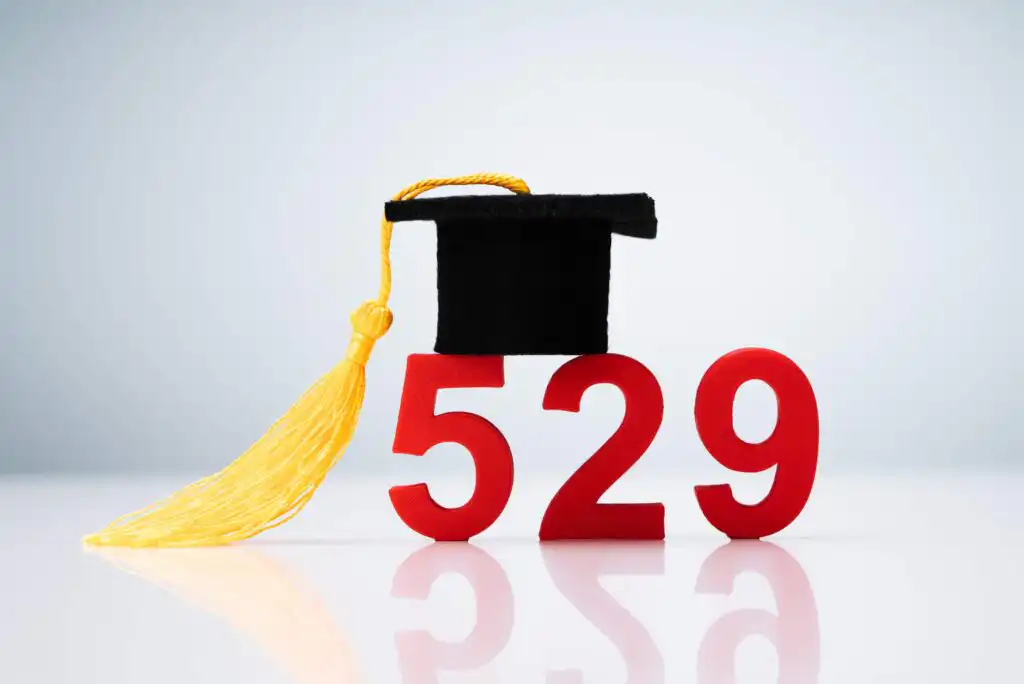 Red numbers “529” with a black graduation cap and yellow tassel resting on top, symbolizing the 529 college savings plan, on a white background with a subtle reflection.