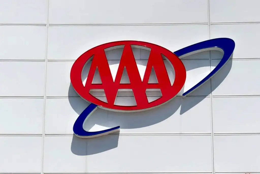 The image shows the AAA (American Automobile Association) logo, featuring three red capital As inside a red oval with a blue swoosh, mounted on a white wall with horizontal panel lines.