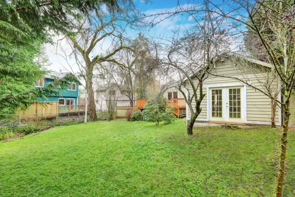 A spacious backyard with green grass, mature trees, and a wooden deck attached to a beige house with French doors; neighboring houses and a wooden fence are visible.