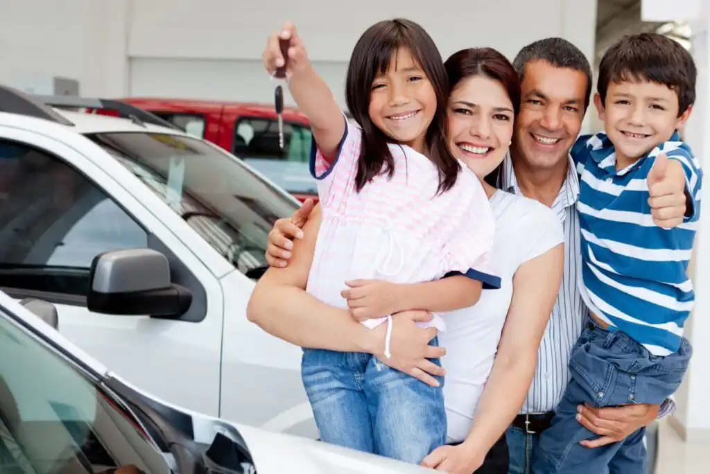 A smiling family of four stands by a white car. The mother hugs a daughter holding car keys, while the father hugs a son who is giving a thumbs up. They appear happy, likely celebrating getting a new car.