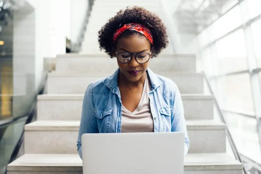 A woman with curly hair and a red headband sits on stairs indoors, focused on her laptop. She wears glasses, a denim shirt, and a neutral top. Natural light streams in from large windows behind her.