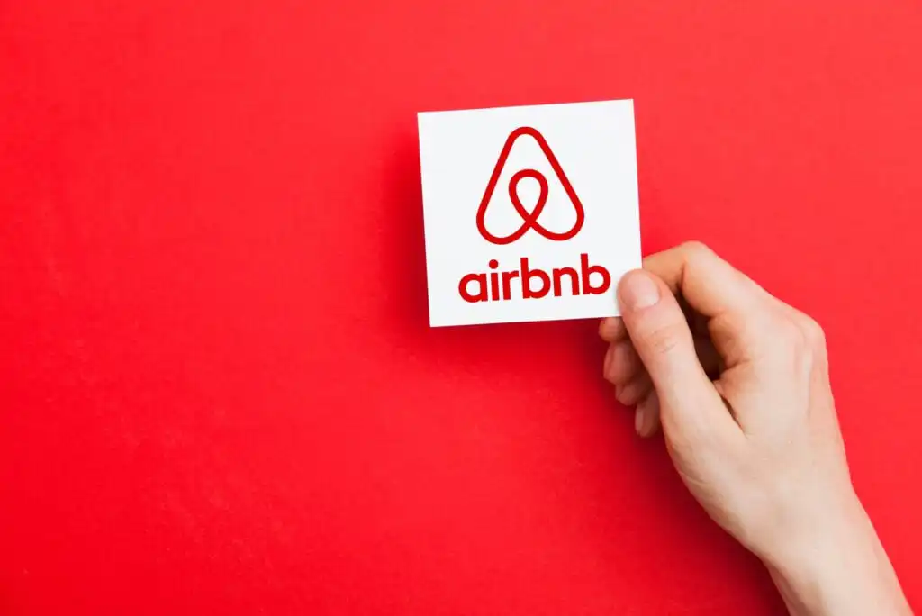 A hand holds a white card with the red Airbnb logo and the word airbnb on a bright red background.
