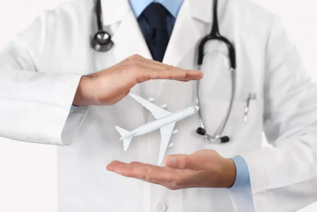 A doctor wearing a white coat and stethoscope holds his hands around a small model airplane, symbolizing medical tourism or healthcare and travel.