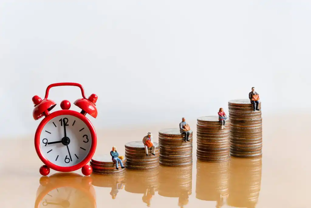 A red alarm clock sits next to ascending stacks of coins, each topped with a miniature figurine, symbolizing people and the growth of money over time.