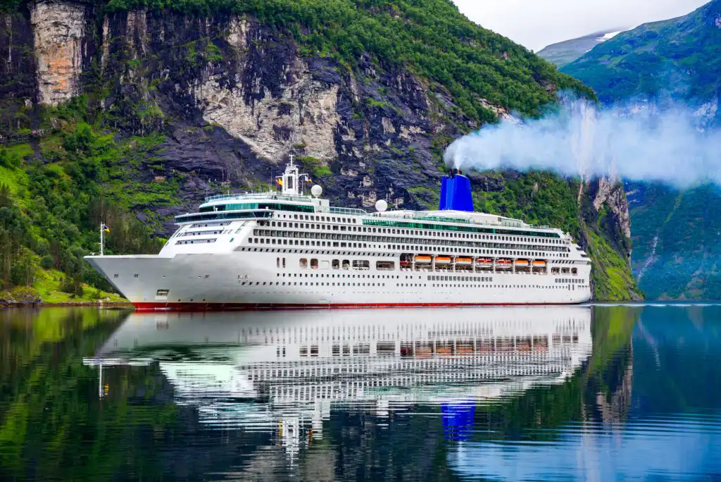 A large white cruise ship with a blue funnel emits smoke as it sails on calm water, reflecting the vessel. Steep, green mountains and rocky cliffs tower in the background under a partly cloudy sky.