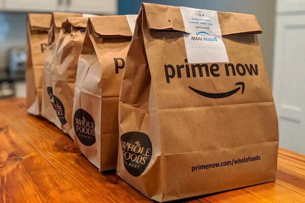 Three brown paper grocery bags from Whole Foods Market with Amazon Prime Now branding are lined up on a wooden table, ready for delivery or pickup.