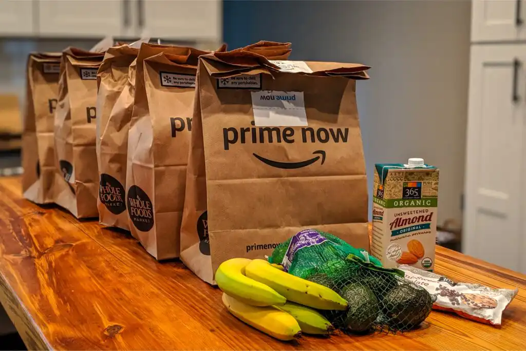 Five brown paper grocery bags labeled Prime Now are lined up on a wooden table. In front of them are bananas, avocados, a carton of almond milk, and a bag of chocolate chips. Kitchen cabinets are in the background.