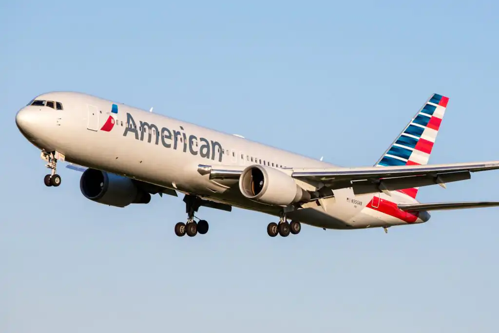 An American Airlines passenger airplane in flight, landing gear extended, flying against a clear blue sky.