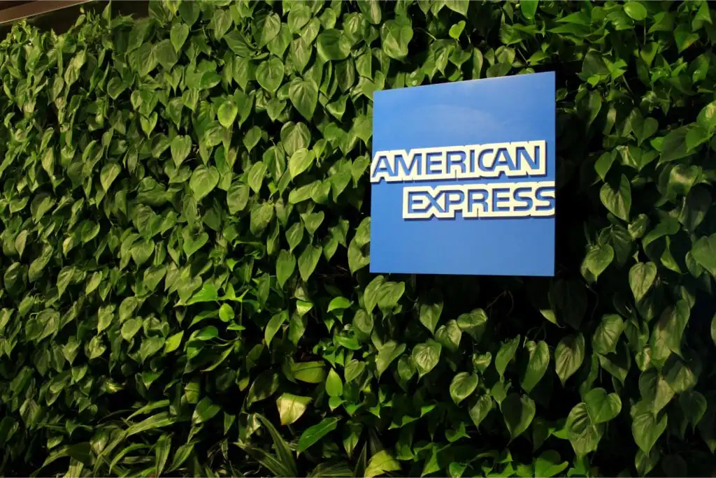 A blue American Express logo sign mounted on a lush green wall covered with heart-shaped leaves.