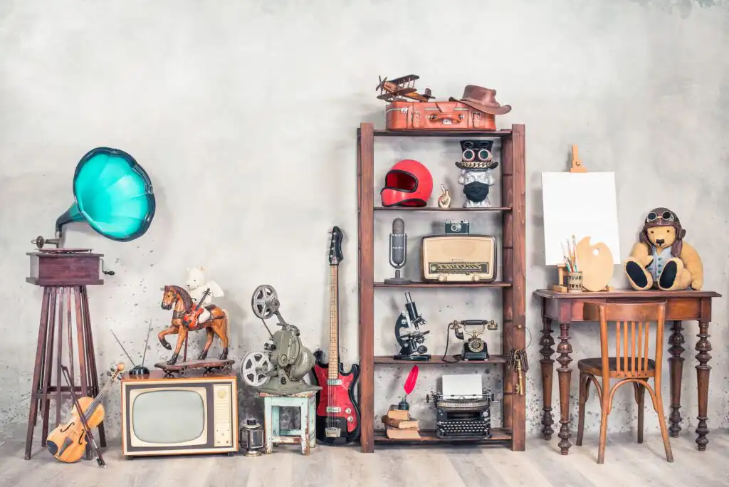 A collection of vintage items including a gramophone, old television, rocking horse, film projector, guitar, typewriter, camera, helmet, and teddy bear arranged on a shelf and tables against a gray wall.