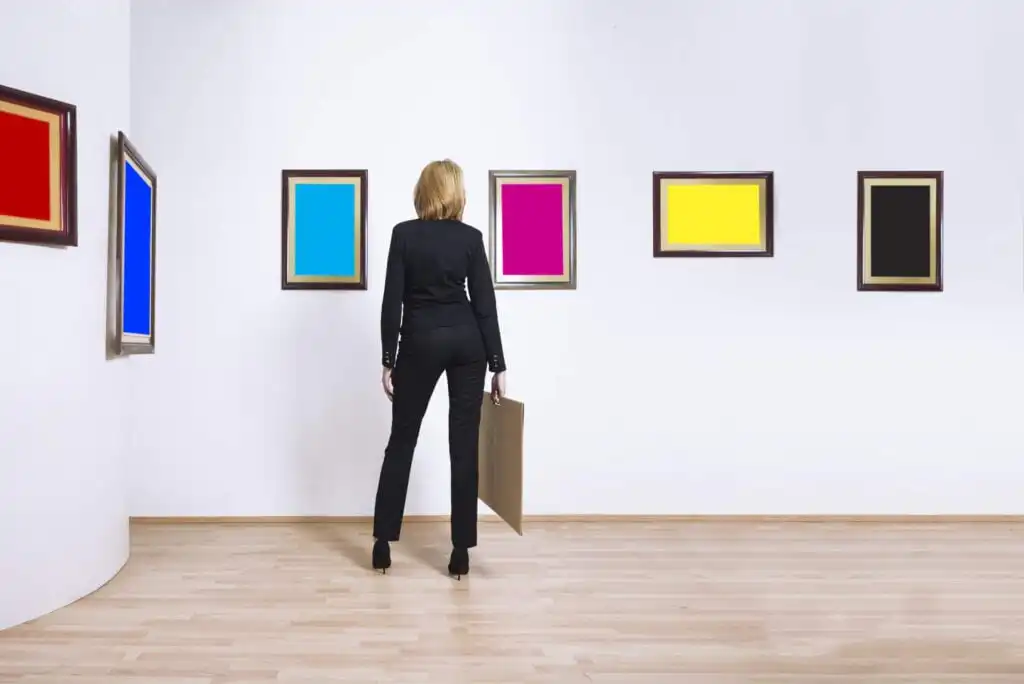 A person in a black suit stands in a gallery, holding a picture frame and looking at three framed rectangles on the wall—colored blue, pink, and yellow—from left to right.
