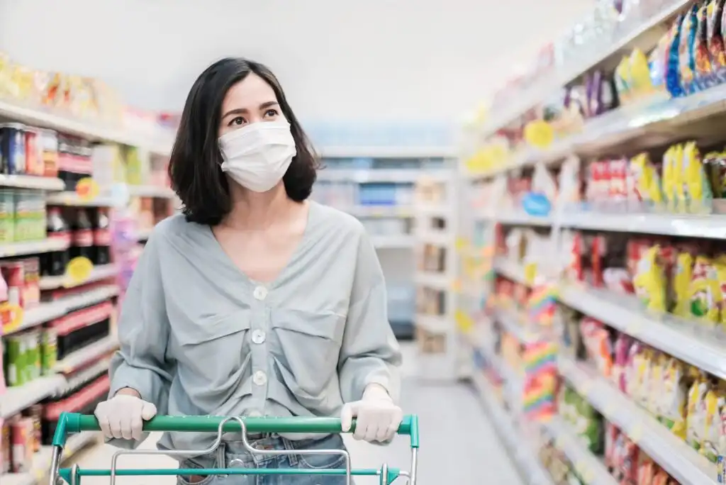 A woman wearing a face mask and gloves pushes a shopping cart down an aisle in a brightly lit grocery store, surrounded by shelves stocked with various products.