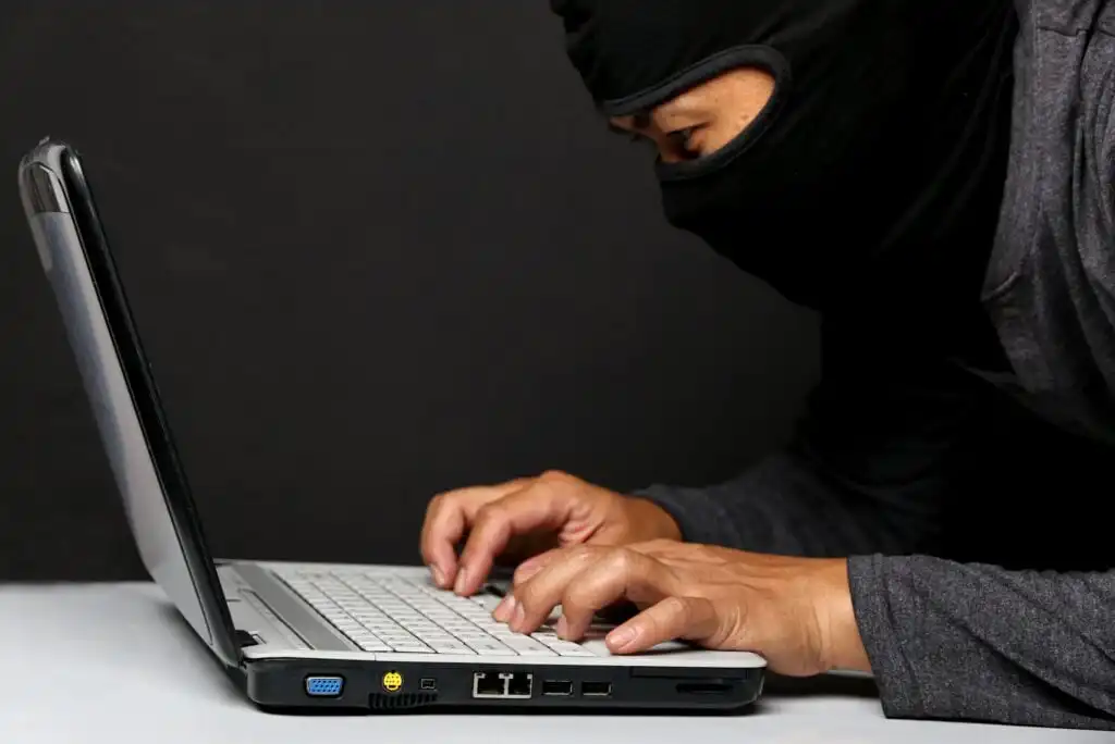 A person wearing a black ski mask and dark clothing is hunched over a laptop, typing on the keyboard against a plain black background, suggesting secretive or suspicious activity.