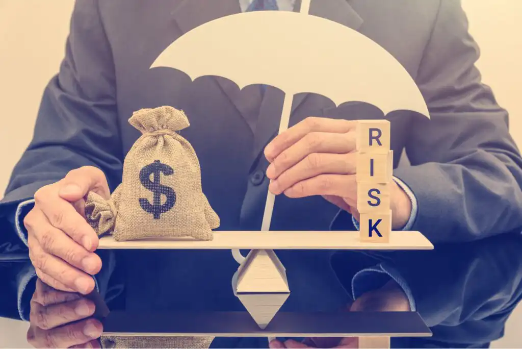 A person in a suit holds a white umbrella over a seesaw balancing a money bag and wooden blocks spelling RISK, symbolizing financial protection and risk management.