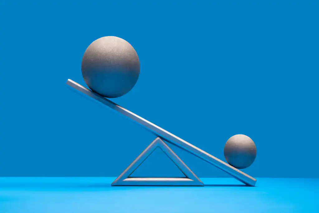 A large metallic sphere and a small metallic sphere sit at opposite ends of a tilted silver balance beam on a triangular base, against a solid blue background.