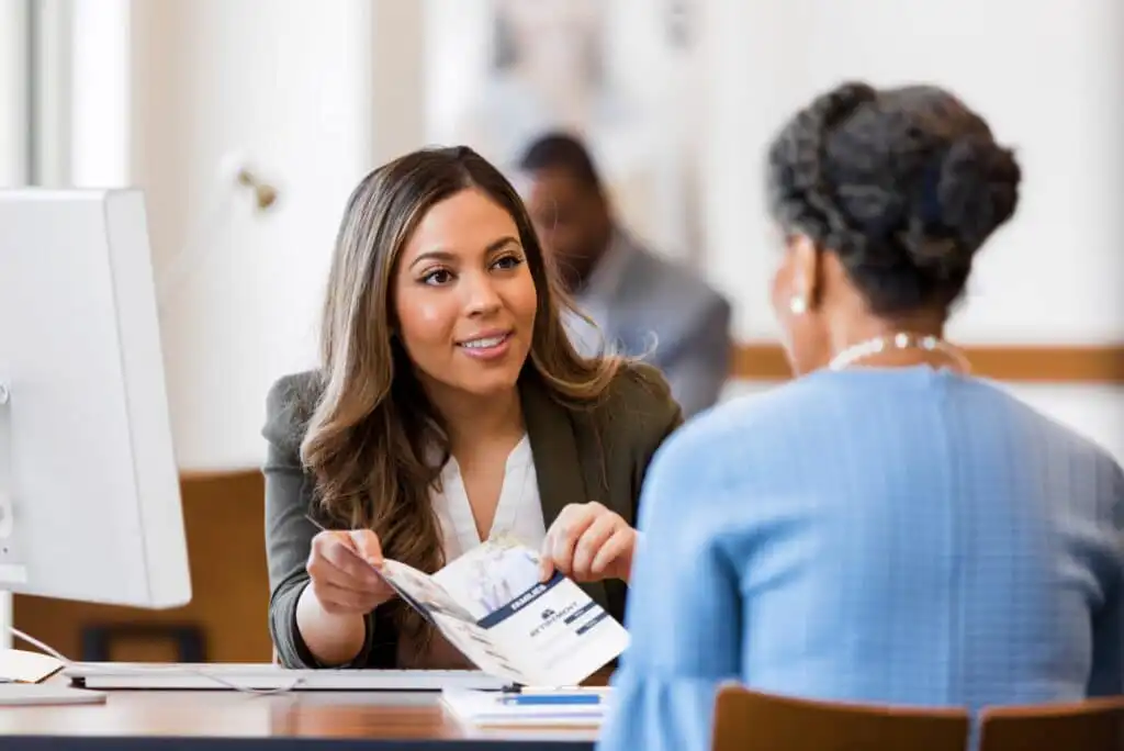 A businesswoman smiles and shows a brochure to another woman across a desk in a modern office setting. A computer monitor and paperwork are visible on the desk.
