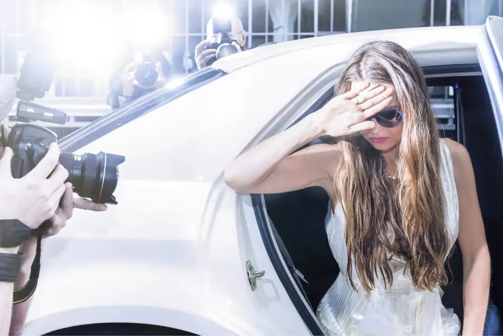 A woman in a white dress and sunglasses steps out of a car, shielding her eyes from bright camera flashes as paparazzi crowd around, taking photos.