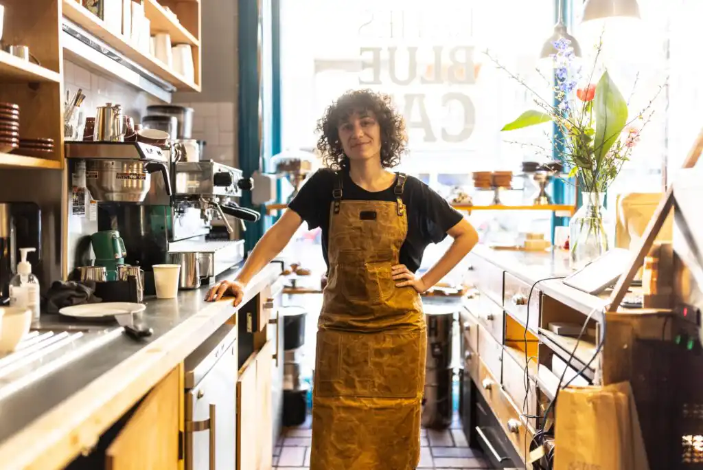 A barista with curly hair, wearing a brown apron over a black shirt, stands confidently behind the counter of a cozy, sunlit café filled with coffee-making equipment and flowers in a vase by the window.