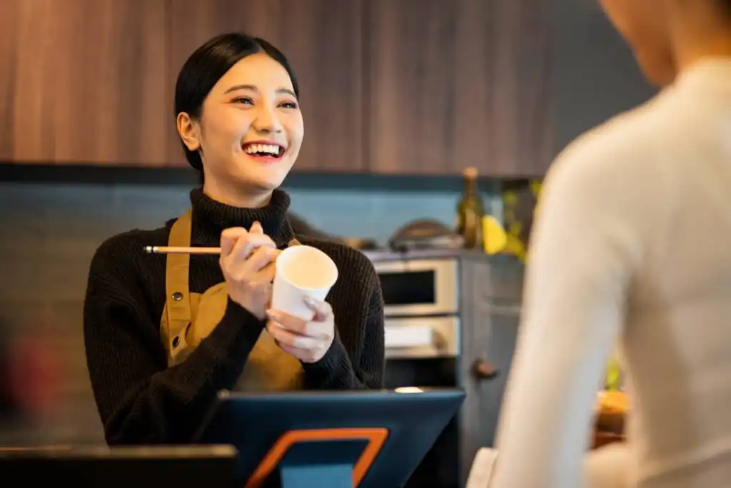 A barista in a dark sweater and apron smiles while holding a notepad and cup, taking an order from a customer at a coffee shop counter.