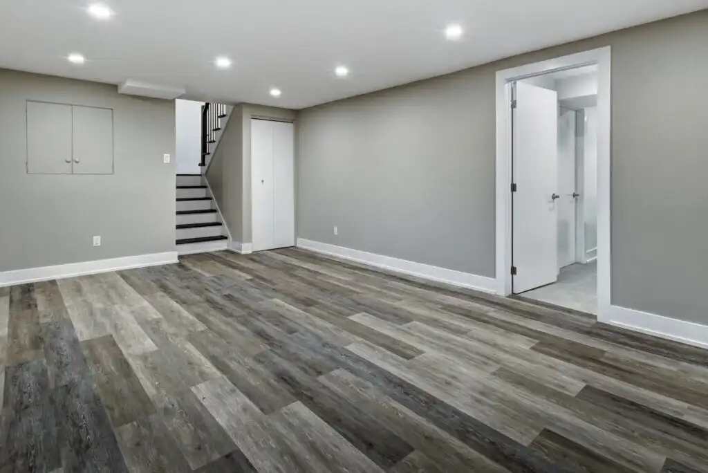A spacious, empty basement room with gray walls, recessed ceiling lights, wood-style vinyl flooring, a closed white door, open white door leading to another room, and a staircase with a black railing.