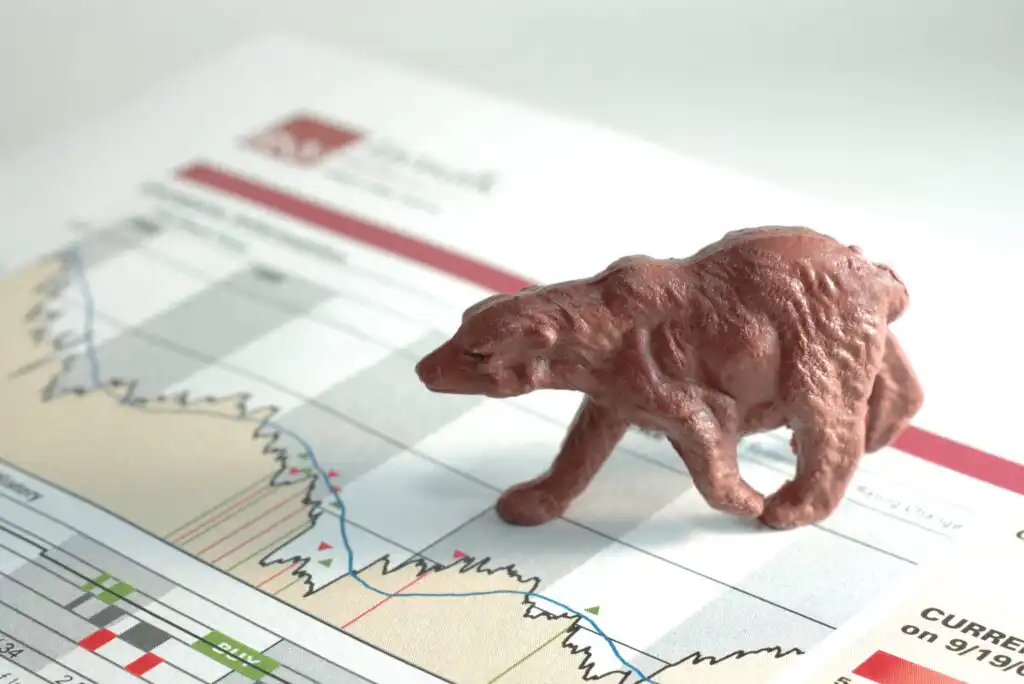 A brown plastic bear figurine is placed on a financial chart with declining lines, symbolizing a bear market or falling stock prices.