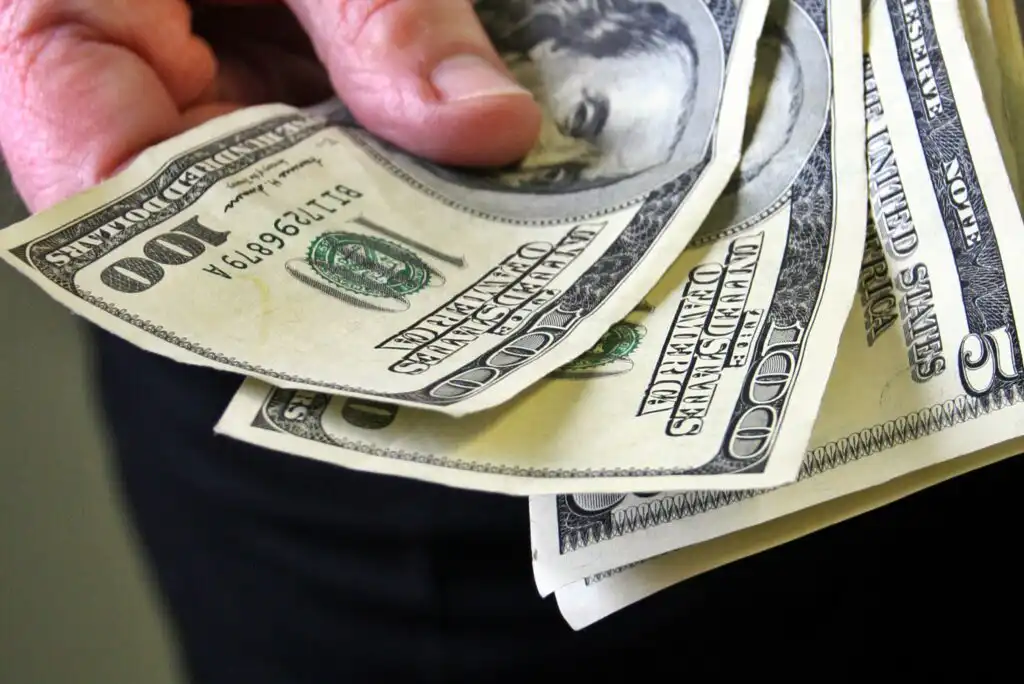 A hand holding several U.S. one hundred dollar bills, fanned out against a dark background.