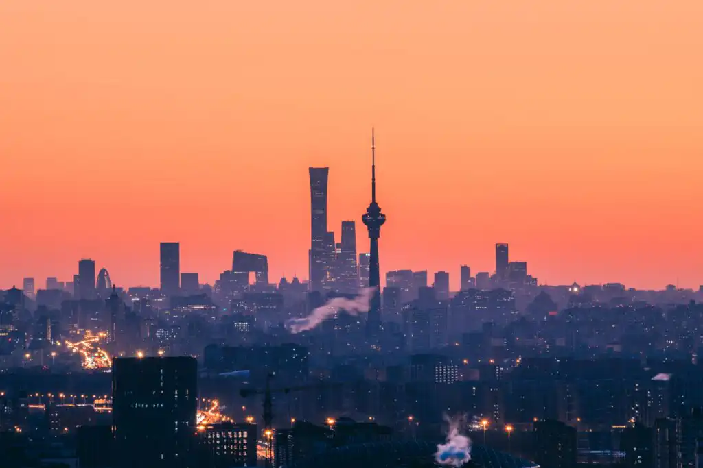 City skyline at sunset with silhouetted skyscrapers and a prominent tower against an orange sky, lights beginning to appear across the city, and wisps of smoke rising in the foreground.