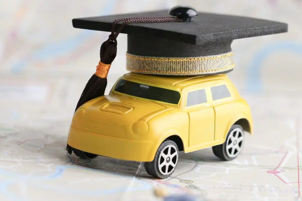 A small yellow toy car with a black graduation cap on top sits on a map background, symbolizing learning or graduation related to driving or travel.