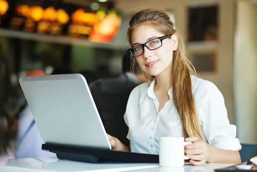 A young woman wearing glasses and a white shirt sits at a table with a laptop, holding a white coffee mug and smiling in a brightly lit café.