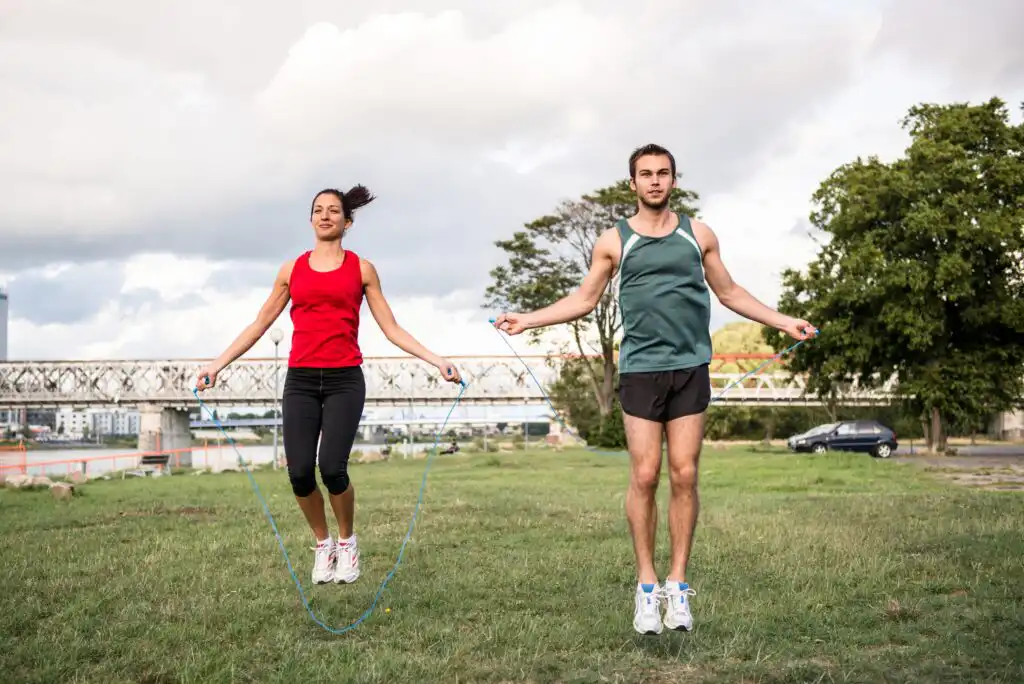 A woman and a man are skipping rope together on a grassy field outdoors. They are both dressed in athletic wear, and a bridge and trees are visible in the background.