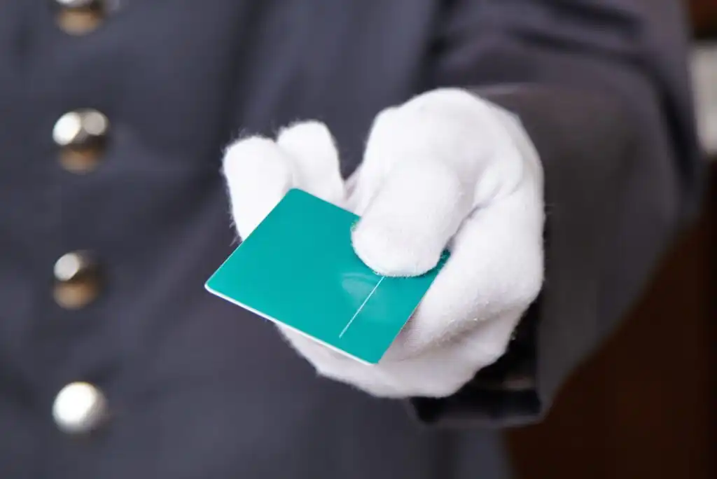 A person wearing a dark uniform and white gloves is holding out a turquoise key card. The focus is on the gloved hand and the card, with the uniforms buttons visible in the background.