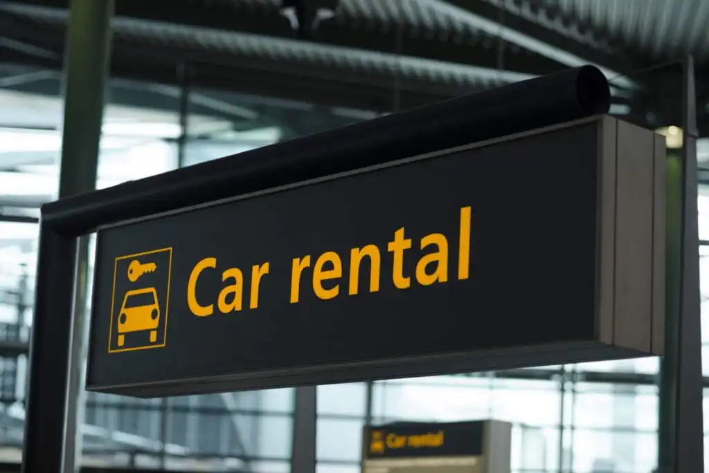 A close-up of a black airport sign with yellow text and icons indicating Car rental, with a background of large windows and structural beams.
