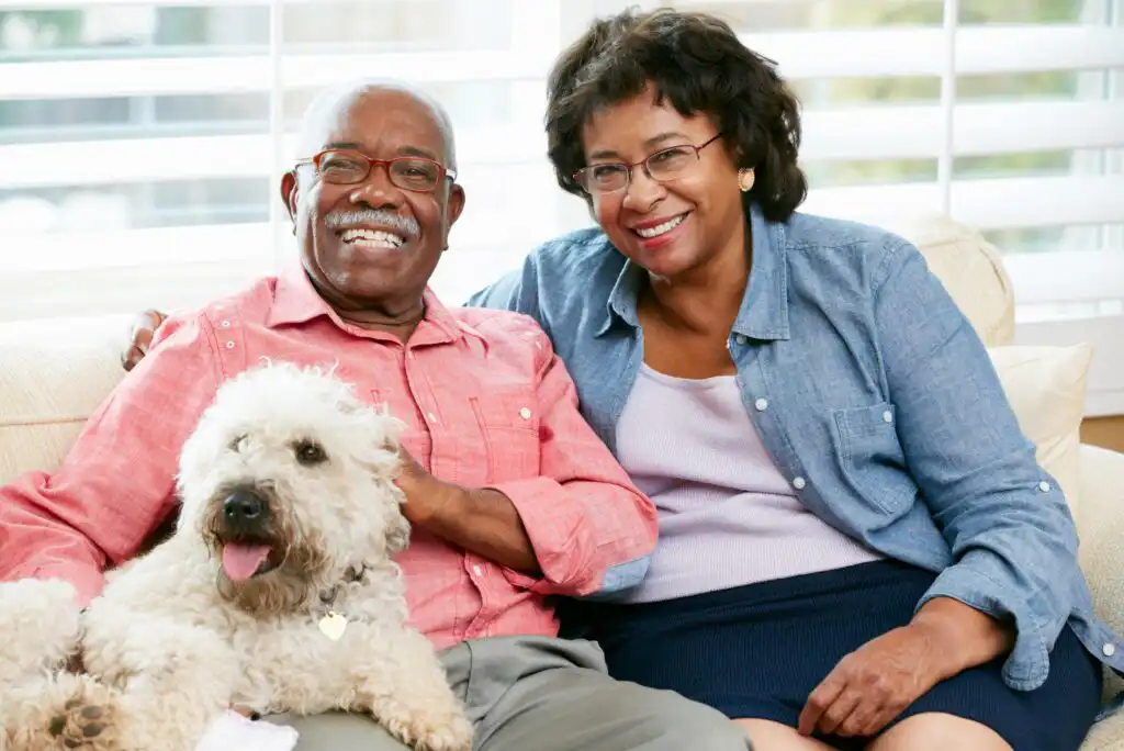 An older couple sits closely together on a couch, smiling at the camera. The man has his arm around a fluffy white dog sitting on his lap. They appear happy and relaxed in a bright, cozy living room.