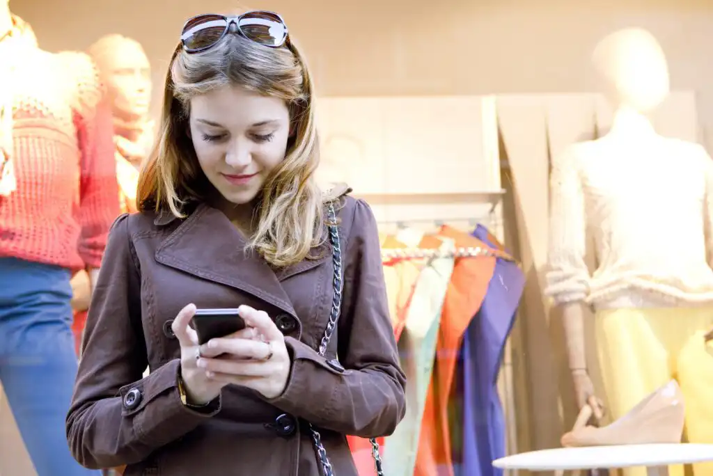 A woman wearing a brown coat and sunglasses on her head looks down at her smartphone while standing in front of a clothing store window with mannequins dressed in colorful outfits.