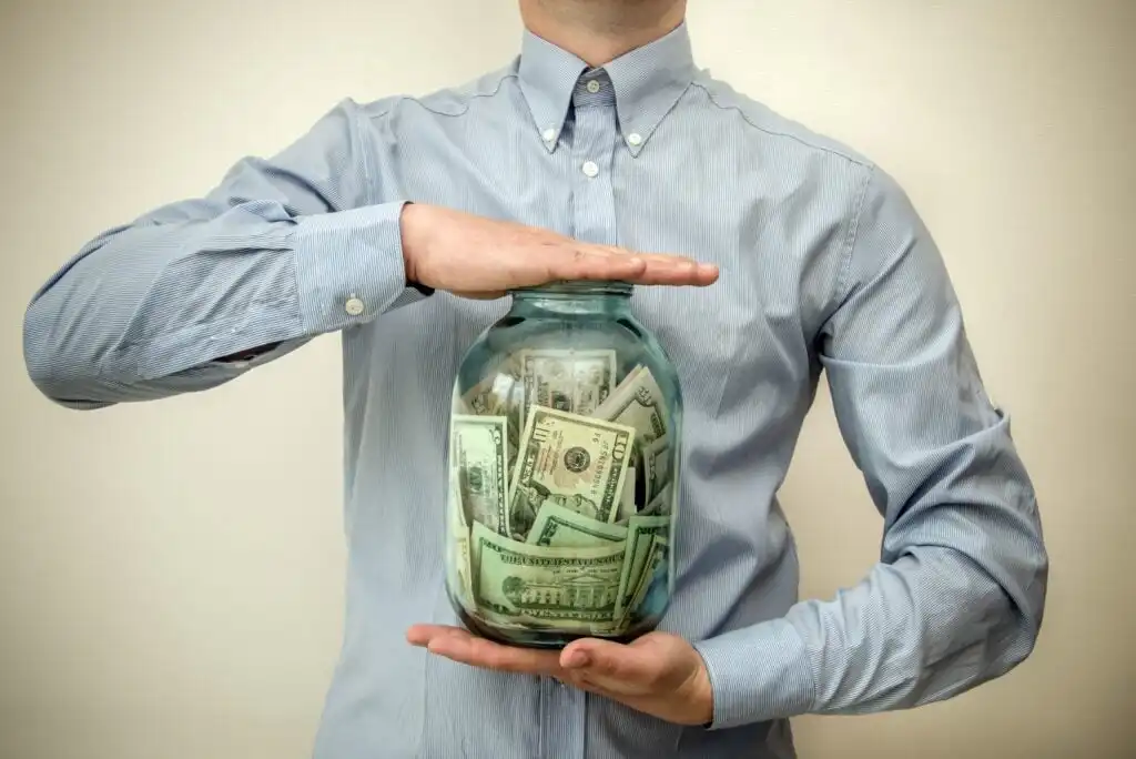 A person in a light blue button-up shirt holds a large glass jar filled with U.S. dollar bills, with one hand supporting the jar and the other resting on top.