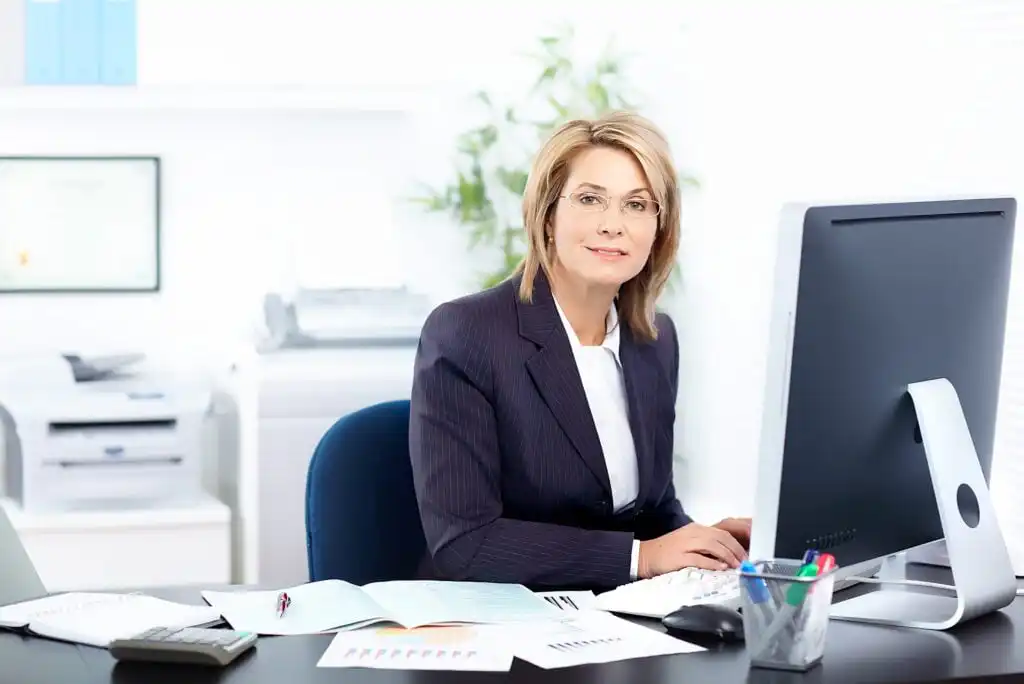 A woman in a business suit sits at a desk in an office, working on a computer. Papers, a calculator, and pen holders are on the desk. She is looking at the camera and smiling slightly. The background is bright and organized.
