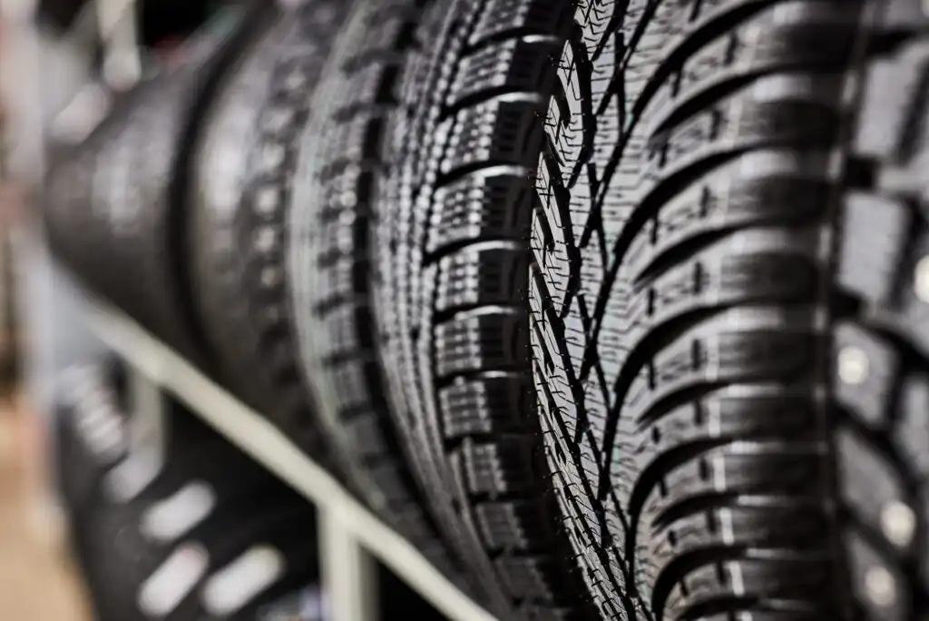 Close-up view of several black car tires with detailed tread patterns, stacked vertically on a metal rack in a store or garage setting.