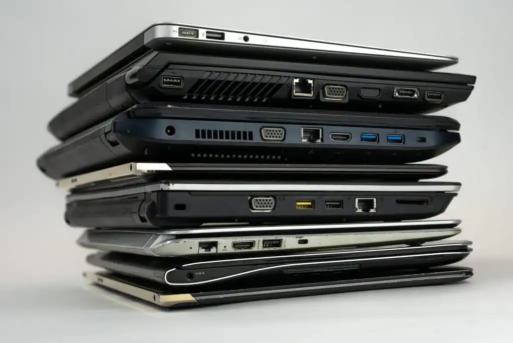 A stack of various closed laptop computers is shown from the side, displaying different ports and thicknesses against a plain background.