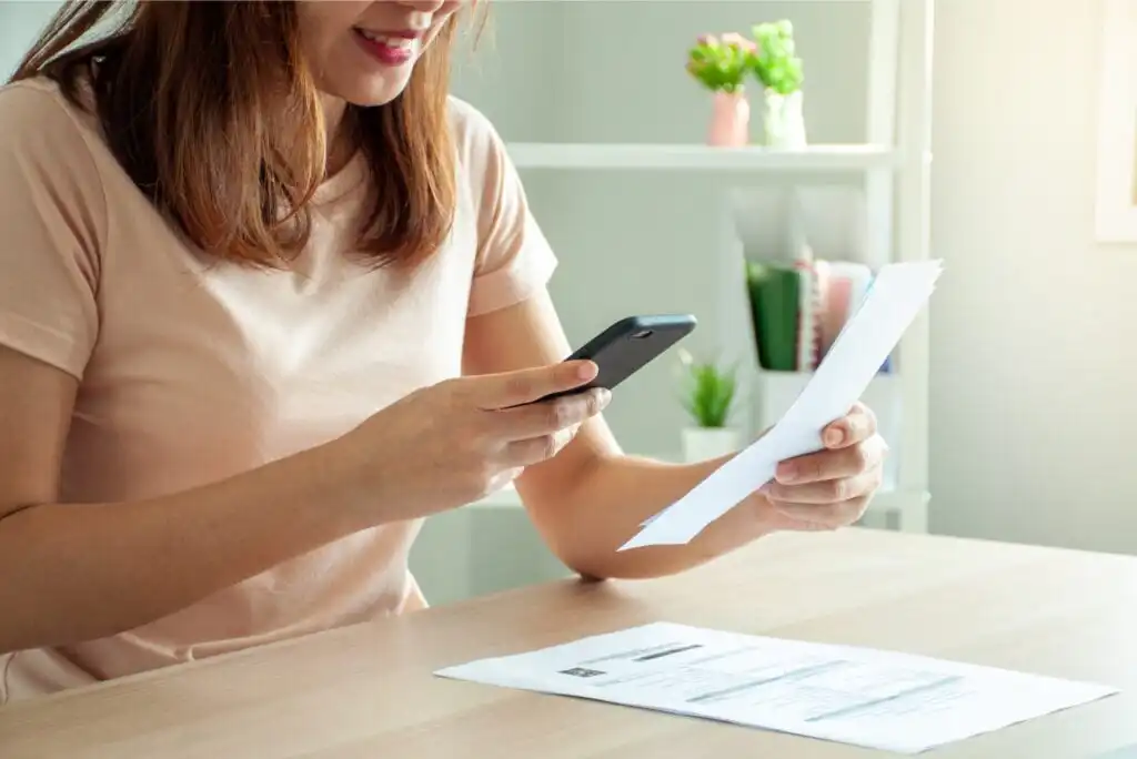 A woman sits at a desk, smiling while using her smartphone to take a photo of a document she is holding. Additional papers are on the table, with a shelf and plants in the background.