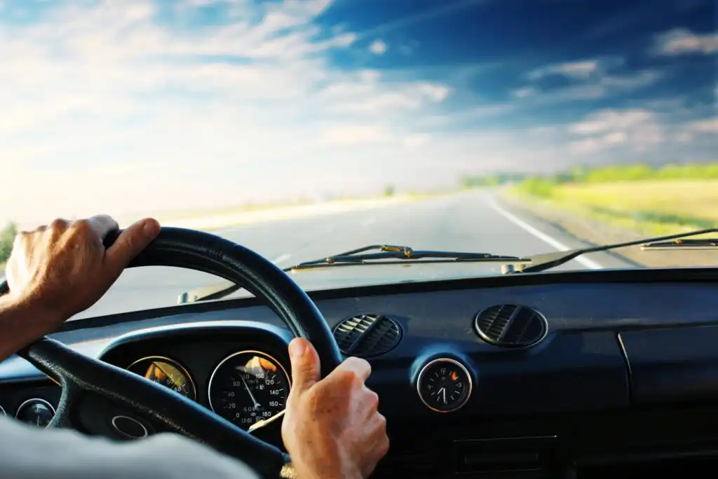 A person’s hands gripping a steering wheel, driving a car down an empty highway with clear blue skies and green fields visible through the windshield. The dashboard and speedometer are also visible.