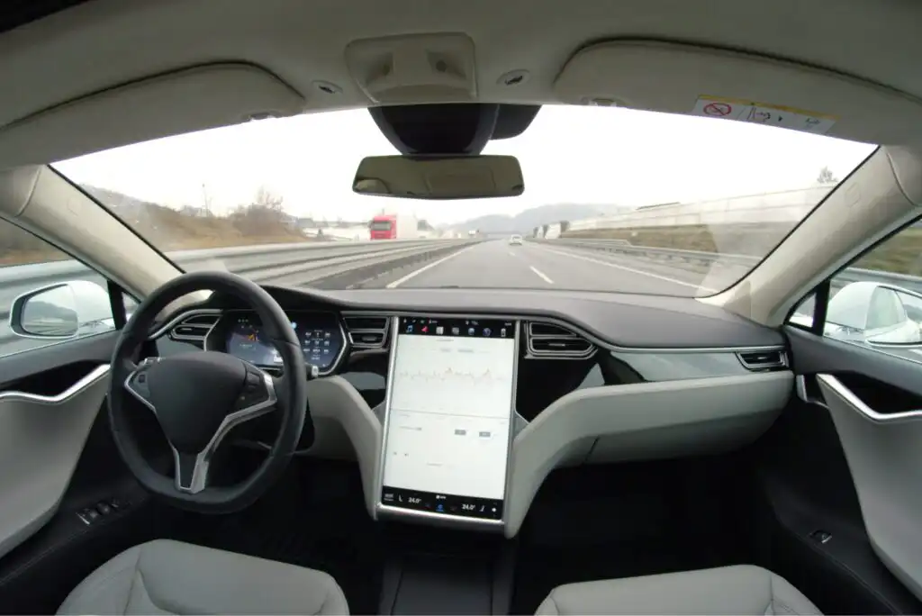 View from inside a modern car driving on a highway, showing the steering wheel, dashboard, and a large central touchscreen display, with no driver visible and vehicles on the road ahead.