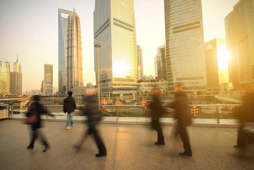 People walk past tall, modern skyscrapers in a city at sunset. The sunlight reflects off the glass buildings, and the scene shows motion blur as people move across a pedestrian bridge.