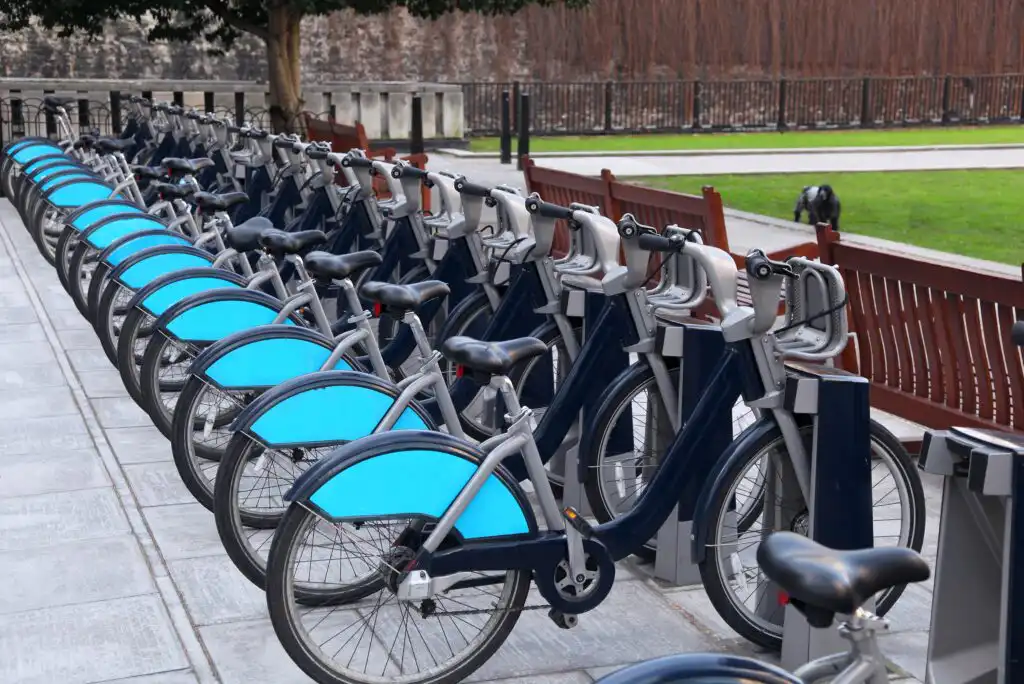 A row of public rental bicycles with turquoise rear wheel covers are parked at a bike sharing station on a paved sidewalk next to a park with grass and benches.