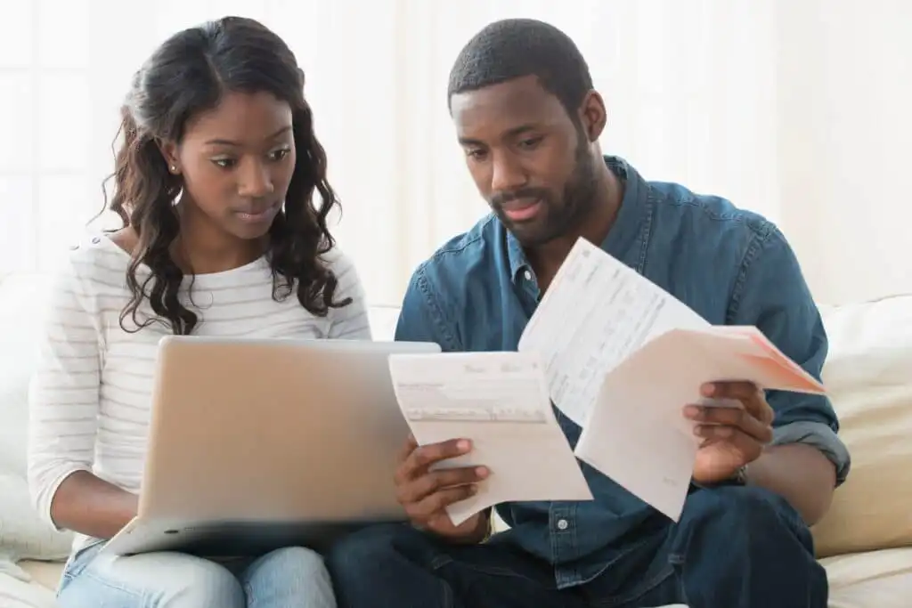 A couple sits together on a couch, looking at a laptop and reviewing several papers and bills, appearing focused and concerned.